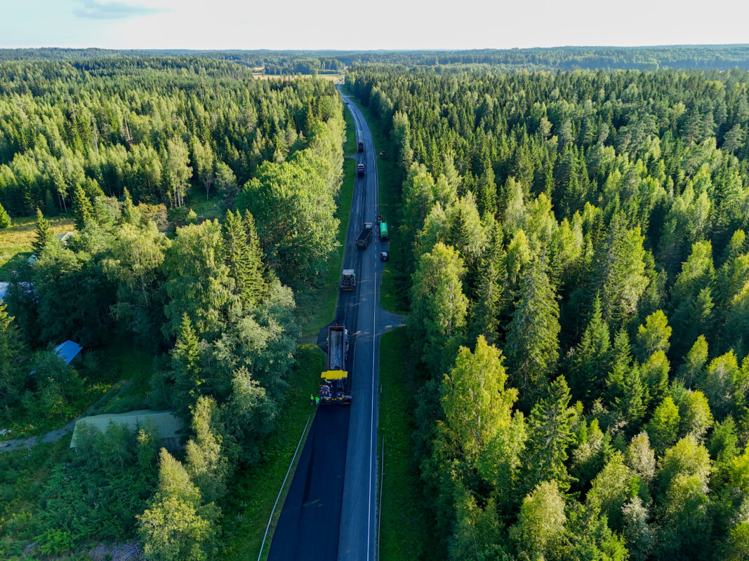 Aurinkoisen metsän läpi kulkevan tien asfaltointia ylhäältä päin kuvattuna.